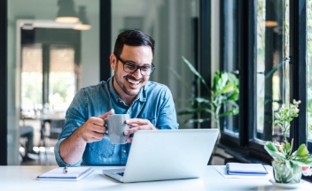 Man in shirt holding a mug looking at laptop.
