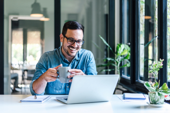 Male in blue shirt sitting at laptop holding a mug.